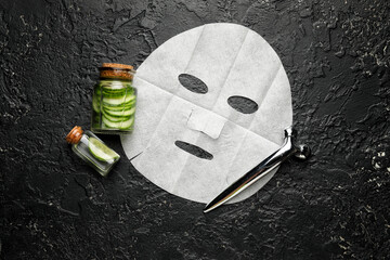Composition with fabric facial mask, massage tool and cucumber essential oil on dark background