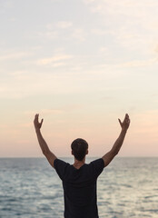 Healthy young man with arms up celebrating during a beautiful sunset facing the ocean. Wellness concept.