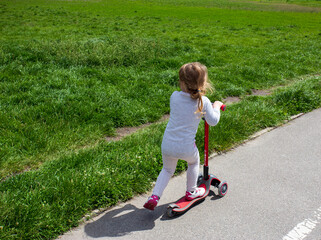 Little blond girl riding a red scooter in the green field © Svetlana