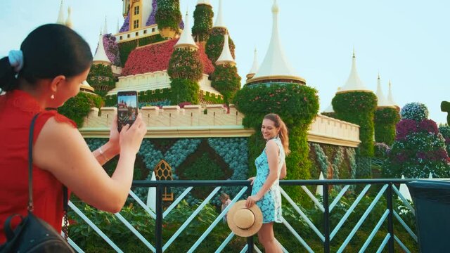 Two women friends on vacation. Happy tourist model posing on backdrop colorful flowering garden. girl photographs lady on mobile phone. Woman taking picture. enjoy beauty garden city Dubai 4k UAE 2020
