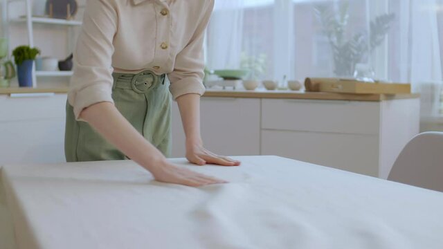 Lockdown of unrecognizable woman wearing casual clothes laying beautiful beige tablecloth on dining table and then smoothing it with hands