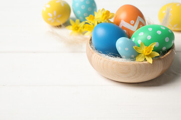 Colorful Easter eggs and flowers in bowl on white wooden table. Space for text