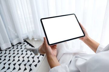 Naklejka premium Mockup image of a woman holding black tablet pc with blank white desktop screen while sitting in bedroom with feeling relaxed in the morning