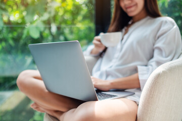 Closeup image of a woman working on laptop computer while drinking coffee and sitting on a white armchair at home with green nature background
