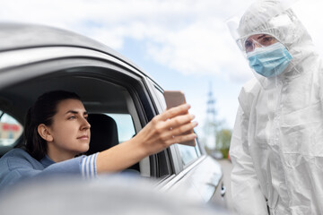 medicine, quarantine and pandemic concept - woman in her car showing her smartphohe to doctor or healthcare worker in protective gear or hazmat suit, medical mask, face shield and goggles