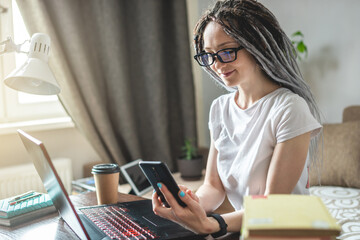 Young modern female employee is working remotely online at home in room using phone and laptop. Distance freelance work