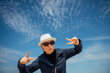 Cheerful woman in white hat and sunglasses poses against beautiful blue sky with feathery clouds. Stylishly dressed girl smiles and gestures with her hands. Happiness, positive, pleasure, joy.