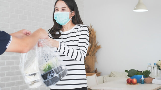 Asian Woman Face Mask At Home And Deliver Man Wearing Face Mask Handling Bag Of Food, Fruit, Vegetable Give To Female Costumer In Front Of The House.  Grocery Delivery Service During Covid19.