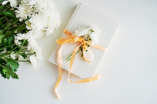 On the table lies a white book tied with a gift ribbon. Nearby lies a bouquet of chrysanthemums