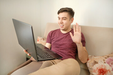 Attractive happy young man, sitting at home, using laptop computer, having video chat, waving hand. Uses video to chat with friends and family.