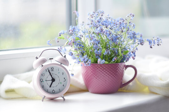 spring bouquet of flowers and a cozy white plaid on the windowsill. pink clock by the window shows morning time