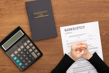 cropped view of female clenched hands on bankruptcy paper near bankruptcy law book and calculator on wooden background