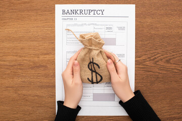 cropped view of woman holding money bag near bankruptcy paper on wooden background