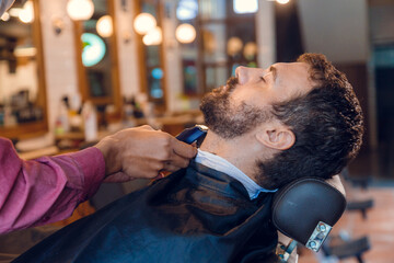 Beard styling. Barber shaping beard of handsome man using a trimmer. Skilled barber trimming beard...