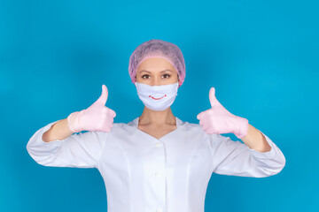 A beautiful doctor in a medical mask, cap and gloves shows a symbolic gesture with his hands with a thumbs up on a blue background. Everything will be alright.  Stop COVID-19. Copy space. Mock up