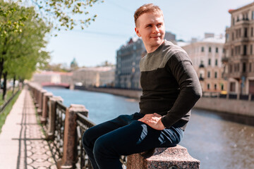 Fototapeta premium A businesslike young man is sitting on the embankment on a Sunny day, green trees everywhere, walking around