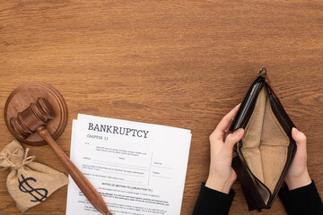 cropped view of woman holding empty wallet near bankruptcy documents, money bag and gavel on wooden...