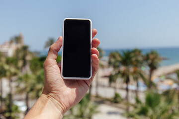 Cropped view of man holding smartphone with palm trees and sea coast at background