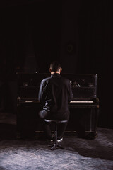 Man in black suit sit in front of piano in black interior, rear view.