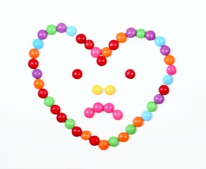 Sad heart made from colored smarties. Chocolate candies in a shape of heart with sadness on face, white background,