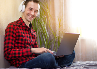 Young man freelancer working with laptop at home in bed near window and listen to music in headphones. Handsome man reads on Internet chronicle of good news from world.
