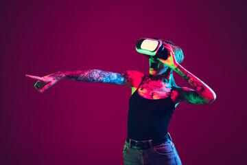 Playing with VR-headset. Young caucasian woman on purple studio background in neon light. Beautiful female model with tattoos. Human emotions, facial expression, sales, ad concept. Freak's culture.