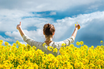  Brunette girl in rapeseed field  summer time, back shot blue sky background.  Happy beautiful young woman on blooming rapeseed field full of joy and happiness. Concept of freedom and childhood