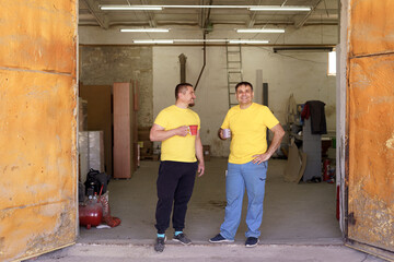 Two carpenters have a tea break while discussing with their workshop in the background