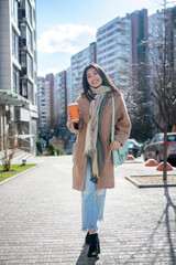 Fototapeta premium Brunette female in beige coat walking along the street with coffee cup, smiling