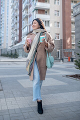 Brunette female in beige coat holding coffee cup, enjoying her drink, walking along the street