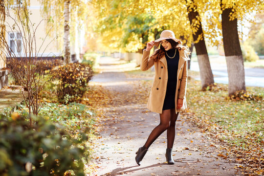 Full length portrait of fashionable woman dressed in beige coat and wide-brimmed hat, dark dress, tights and high hill shoes outdoors in autumn. Yellow leaves on he trees in the background. 