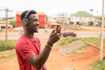 young black man standing outside, using his wireless headphone to listen to songs on his phone