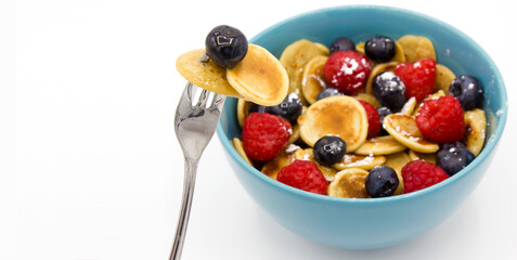 A metal fork with Stack of mini tiny pancakes and berry near blue bowl with heap of cereal mini pancakes . Trend 2020. Home made long breakfast during quarantine. White background. Space for text