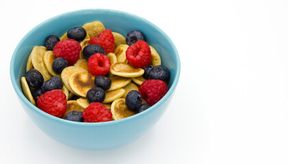 Pancake cereal, mini pancakes with fresh raspberry and blueberry in a blue bowl on white background. Space for text. Side view. Trendy healthy homemade breakfast