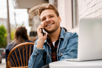 Handsome smiling happy young man hipster in a street cafe talking on a cell phone while working on a laptop. Modern technologies, online communication and internet connection. People lifestyle