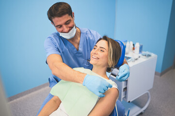 Obraz premium Dentist examining a patient's teeth using dental equipment in dentistry office. Stomatology and health care concept. Young handsome male doctor in disposable medical facial mask, smiling happy woman.