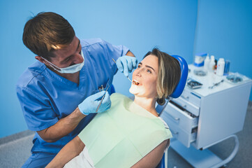 Dentist examining a patient's teeth using dental equipment in dentistry office. Stomatology and health care concept. Young handsome male doctor in disposable medical facial mask, smiling happy woman.