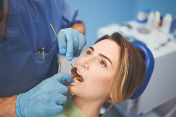 Fototapeta premium Dentist examining a patient's teeth using dental equipment in dentistry office. Stomatology and health care concept. Young handsome male doctor in disposable medical facial mask, smiling happy woman.
