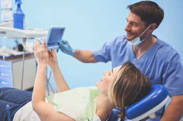 Obraz premium Dentist shows the patient the results of treatment with a mirror, examinating teeth with dental equipment in dental office. Young handsome male doctor in medical facial mask and smiling happy woman.