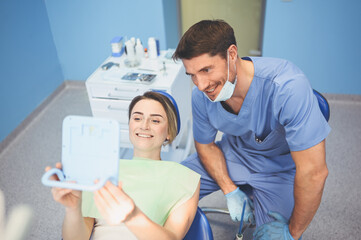 Dentist shows the patient the results of treatment with a mirror, examinating teeth with dental equipment in dental office. Young handsome male doctor in medical facial mask and smiling happy woman.
