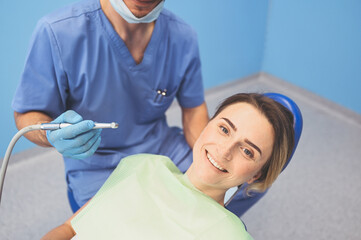 Dentist examining a patient's teeth using dental equipment in dentistry office. Stomatology and health care concept. Young handsome male doctor in disposable medical facial mask, smiling happy woman.