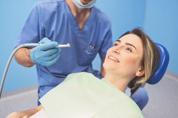 Dentist examining a patient's teeth using dental equipment in dentistry office. Stomatology and health care concept. Young handsome male doctor in disposable medical facial mask, smiling happy woman.