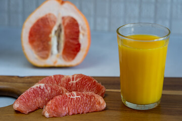 Segments of a grapefruit and a glass of juice standing on wooden cutting board against background of grapefruit half
