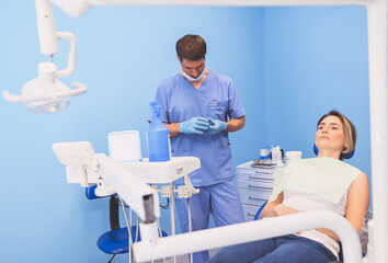 Fototapeta premium Dentist examining a patient's teeth using dental equipment in dentistry office. Stomatology and health care concept. Young handsome male doctor in disposable medical facial mask, smiling happy woman.