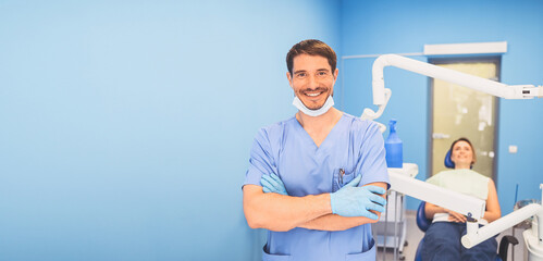 Obraz premium Young smiling handsome male doctor in blue medical uniform, disposable medical facial mask with equipment in dentistry office and happy patient in the dental chair. Stomatology concept. Copy space