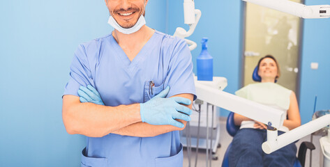Young smiling handsome male doctor in blue medical uniform, disposable medical facial mask with equipment in dentistry office and happy patient in the dental chair. Stomatology concept. Copy space