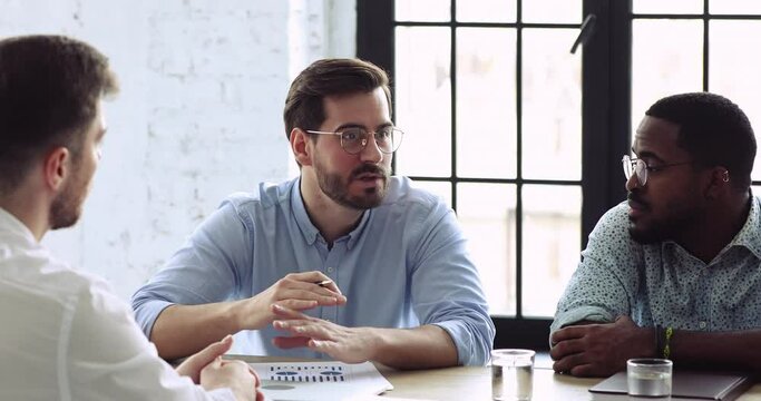 Focused young caucasian male marketing specialist in glasses sharing research results to focused multiracial colleagues at briefing meeting. Smart diverse board members summarizing working results.