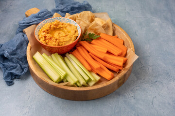 Bowl of butternut squash hummus on a wooden tray with carrots, celery, and pita bread against a blue textured background