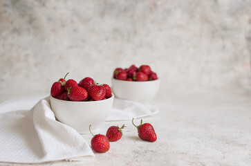 Strawberries in a white ceramic bowl on the table. Nearby lies a white textile towel and several red berries. In the background is another bowl of strawberries. Selective focus.