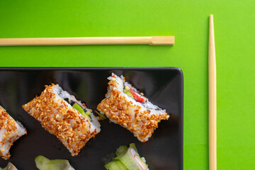 Top down view of a black square plate with multiple sushi rolls with sesame seeds and cucumber on green simple background with chopsticks paralel to the plate